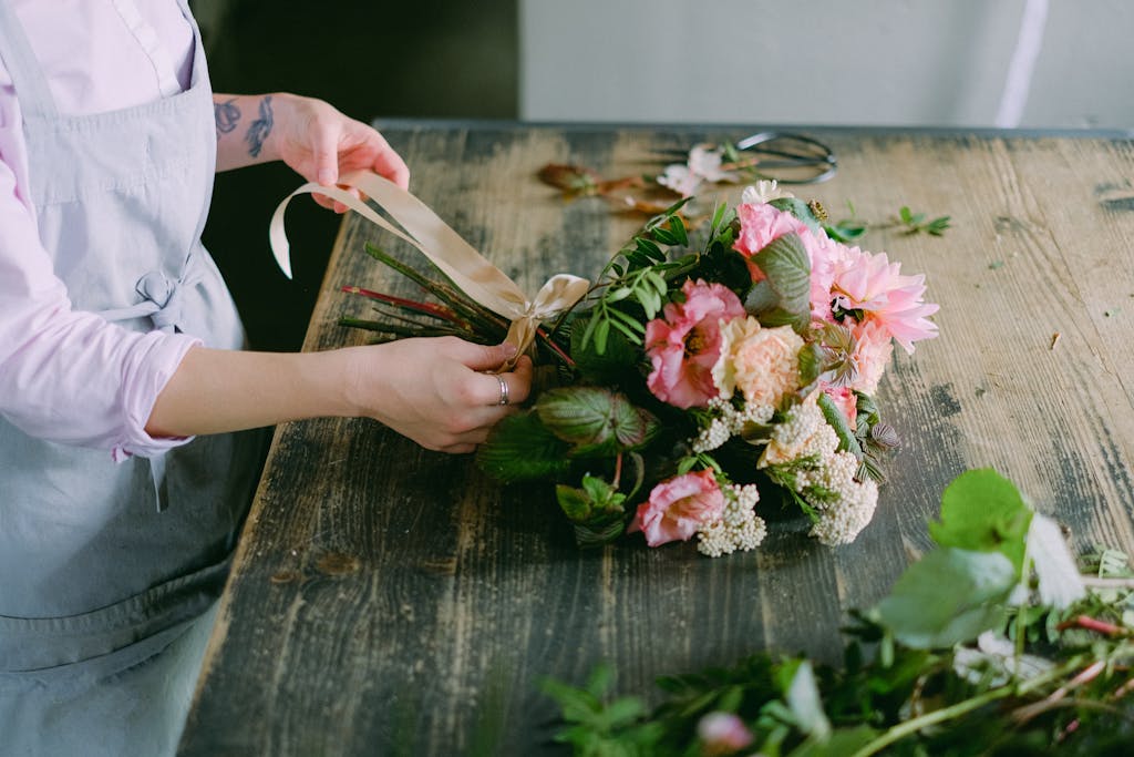 Florist tying ribbon on a vibrant flower bouquet on a rustic wooden table indoors.