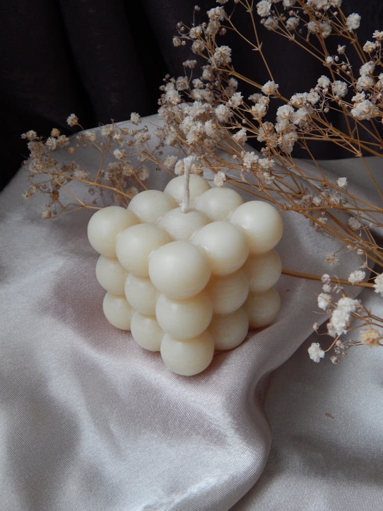 A unique bubble-shaped candle surrounded by dried flowers on a silky fabric backdrop.